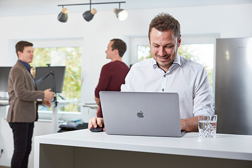 Nico Wohlleb programmiert am Laptop während zwei Personen im Hintergrund sprechen